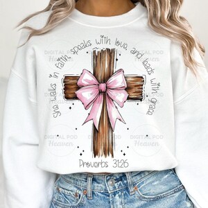 May include: White sweatshirt featuring a brown wooden cross with a pink bow. The cross has the text "She walks in faith, speaks with love, and leads with grace" and "Proverbs 31:26" printed on it.