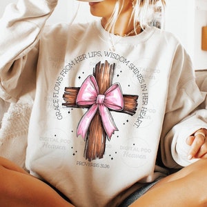 May include: A beige sweatshirt featuring a brown wooden cross with a pink bow. The cross is encircled by the text "GRACE FLOWS FROM HER LIPS, WISDOM SHINES IN HER HEART" and "PROVERBS 3:26".