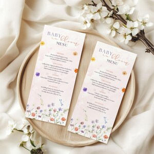 May include: Two pink baby shower menus with floral designs and the text "BABY in BLOOM MENU" are displayed on a wooden tray. The menus feature a list of courses and beverages, with colorful flower illustrations.