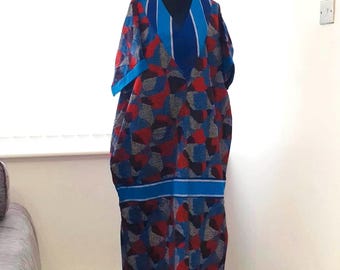 May include: A long, flowing dress with a V-neck and short sleeves. The dress features a vibrant pattern of red, blue, and gray geometric shapes on a dark background. Blue accents highlight the neckline and waist. The dress is displayed on a black mannequin.