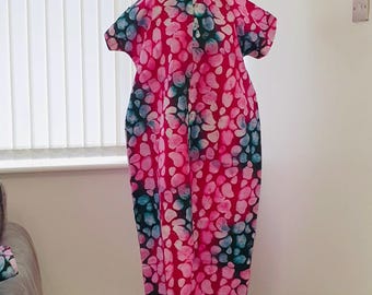 May include: A long, flowing dress with a vibrant pattern of pink, blue, and teal organic shapes against a red background. The dress has short sleeves and a mandarin collar, buttoned down the front. The garment is displayed on a black mannequin.