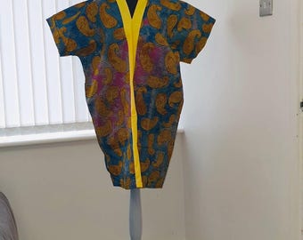 May include: A colorful kimono-style garment with a paisley pattern in shades of blue, yellow, and pink. The kimono has a yellow trim and is displayed on a gray mannequin stand. The garment is made of a patterned fabric.