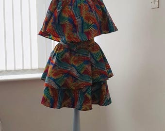 May include: A vibrant, two-piece dress displayed on a mannequin. The top features a cape-like design, and the tiered skirt adds volume. The fabric showcases a colorful, abstract pattern with shades of red, blue, yellow, and green. The dress is a statement piece.