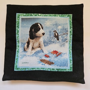 May include: A black throw pillow with a green border features a printed image of a puppy in a snowy winter scene. The puppy is white and black with brown eyes. A small bird is perched on a branch in the background. The image is signed "Giordano".