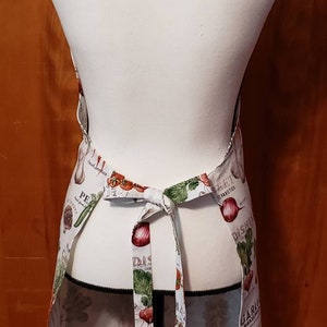 May include: A white apron with a colourful vegetable print. The apron has adjustable ties and features a variety of vegetables, including tomatoes, onions, garlic, and peppers. The text "Fresh Produce" and "Garden Delights" is printed on the apron.