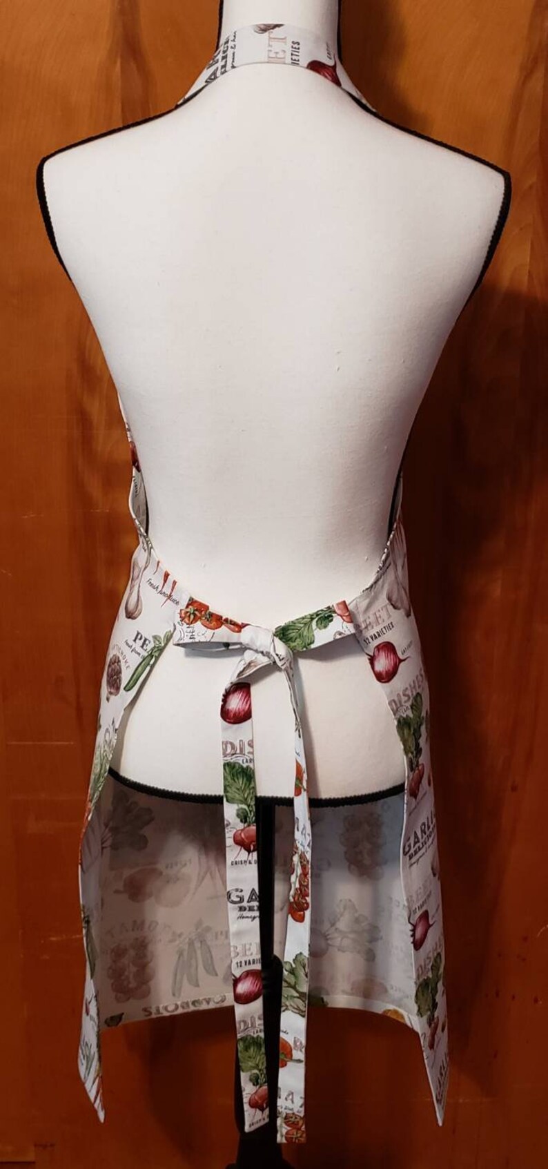 May include: A white apron with a colourful vegetable print. The apron has adjustable ties and features a variety of vegetables, including tomatoes, onions, garlic, and peppers. The text "Fresh Produce" and "Garden Delights" is printed on the apron.
