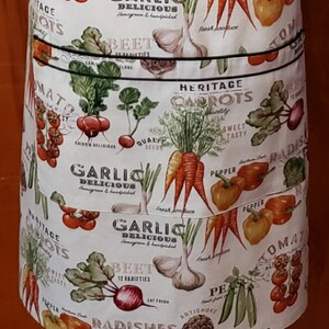 May include: White cotton apron with a colourful print of various vegetables, including carrots, tomatoes, garlic, radishes, and beetroot. The apron has three pockets and features the text "Heritage Carrots", "Garlic Delicious", "Beet 12 Varieties", and "Radishes".