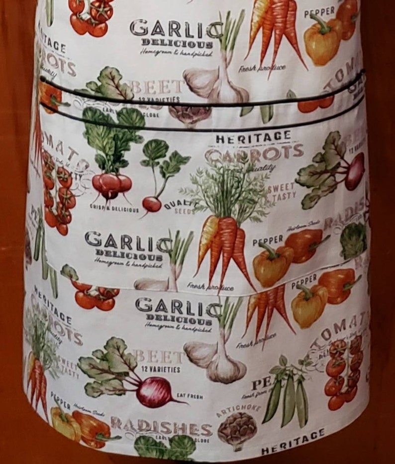 May include: White cotton apron with a colourful print of various vegetables, including carrots, tomatoes, garlic, radishes, and beetroot. The apron has three pockets and features the text "Heritage Carrots", "Garlic Delicious", "Beet 12 Varieties", and "Radishes".