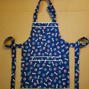 May include: A blue apron with a white trim and a paw print and bone pattern. The apron has two long ties and a large pocket.