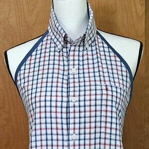 May include: A sleeveless halter-style shirt featuring a white, navy blue, and red plaid pattern. The garment has a button-down front with a pointed collar, dark blue trim along the armholes, and a single chest pocket with a small red logo.