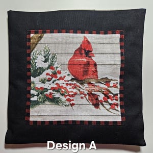 May include: Square black pillow with a central design featuring a red cardinal perched on a snow-covered branch with red berries. The design is set against a gray, wood-grain background and framed by a red and black plaid border. The text "Design A" is at the bottom.