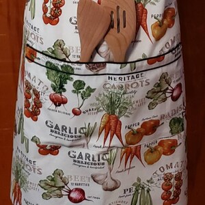 May include: A white cotton apron with a black trim and a large pocket. The apron is covered in a colourful pattern of vegetables, including carrots, tomatoes, radishes, garlic, and beetroot. The text "Heritage Carrots" is printed on the apron.