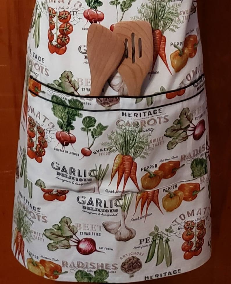 May include: A white cotton apron with a black trim and a large pocket. The apron is covered in a colourful pattern of vegetables, including carrots, tomatoes, radishes, garlic, and beetroot. The text "Heritage Carrots" is printed on the apron.