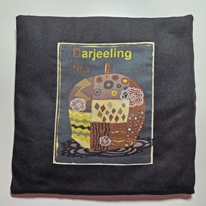 Puede incluir: Cojín cuadrado negro con un diseño de té Darjeeling. El diseño presenta una colorida manzana de estilo patchwork con las palabras "Darjeeling tea" encima. La manzana tiene varios patrones y colores, incluyendo amarillo, marrón y blanco.