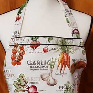 May include: White kitchen apron with a colourful vegetable print. The apron features illustrations of tomatoes, carrots, garlic, radishes, beets, and artichokes. The text on the apron reads "Garlic Delicious Homegrown & Handpicked", "Beet 12 Varieties", "Radishes", and "Fresh Produce".