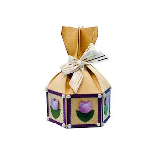 以下が含まれることがあります： 茶色の紙の外側に紫色の縁取りが施された六角形のギフトボックス。ボックスはピンクと緑の紙のチューリップで飾られており、上部に金色のストライプが入った白いリボンが結ばれています。