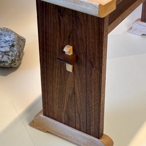 May include: Close-up of a wooden table leg. The leg is made of dark brown wood with a lighter wood accent at the top and bottom. A small, decorative wooden element is attached to the leg. The table appears to be handcrafted.