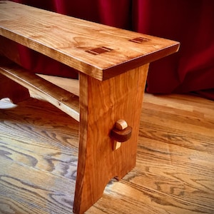 May include: A wooden bench with a smooth, polished finish and visible grain. The rectangular seat features exposed joinery details, while the side support includes a protruding wooden peg fastener. The bench has a sturdy, low-profile design.