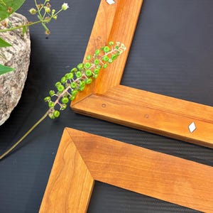 May include: Two wooden picture frames with a natural wood finish, featuring decorative diamond-shaped inlays. A sprig of green berries and white flowers rests near the frames, with a stone vase in the background. The frames are set against a dark background.