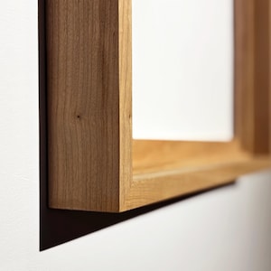 May include: Close-up of a rectangular wooden frame, showcasing the natural grain and warm tones of the wood. The frame is mounted on a white wall, with a shadow cast below. The design is simple and modern, suitable for displaying art or photos.