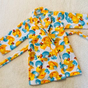 Puede incluir: Bata de baño infantil con un estampado de patos amarillos y burbujas azules sobre fondo blanco. La bata tiene un cinturón y bolsillos a juego. Fabricada con material suave.