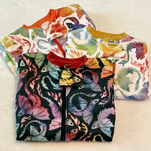 May include: Three colourful zippered jackets with dragon designs. The jackets are made of soft, comfortable fabric and feature a variety of colours, including orange, yellow, blue, and green.