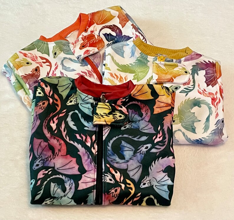 May include: Three colourful zippered jackets with dragon designs. The jackets are made of soft, comfortable fabric and feature a variety of colours, including orange, yellow, blue, and green.