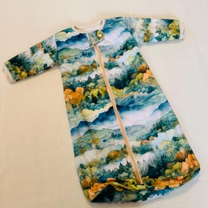 May include: A baby sleep sack with a watercolor mountain scene in shades of blue, green, and orange. The sleep sack has long sleeves, a zipper down the front, and a button closure at the neckline. The sleep sack is designed for infants.