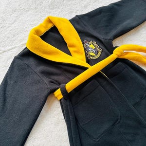 May include: Black fleece robe with a yellow trim and a Hogwarts house crest embroidered on the front. The robe has a matching yellow tie belt.