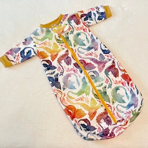 May include: A white baby sleeping bag with a colourful dragon print. The sleeping bag has a yellow zip down the front.