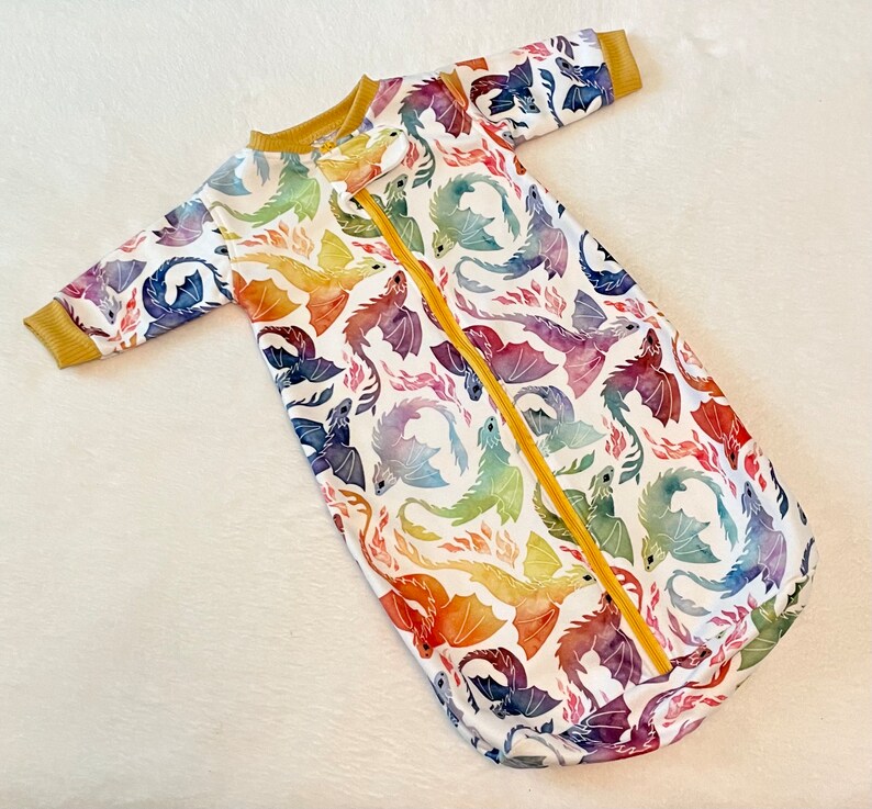 May include: A white baby sleeping bag with a colourful dragon print. The sleeping bag has a yellow zip down the front.