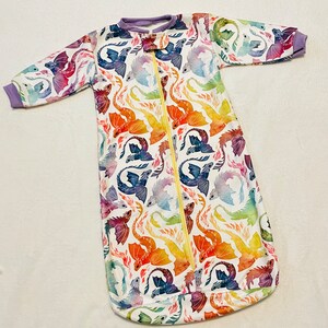 May include: A white baby sleeping bag with a zip fastening and a colourful watercolour dragon print. The dragons are in shades of blue, purple, orange, red and yellow.