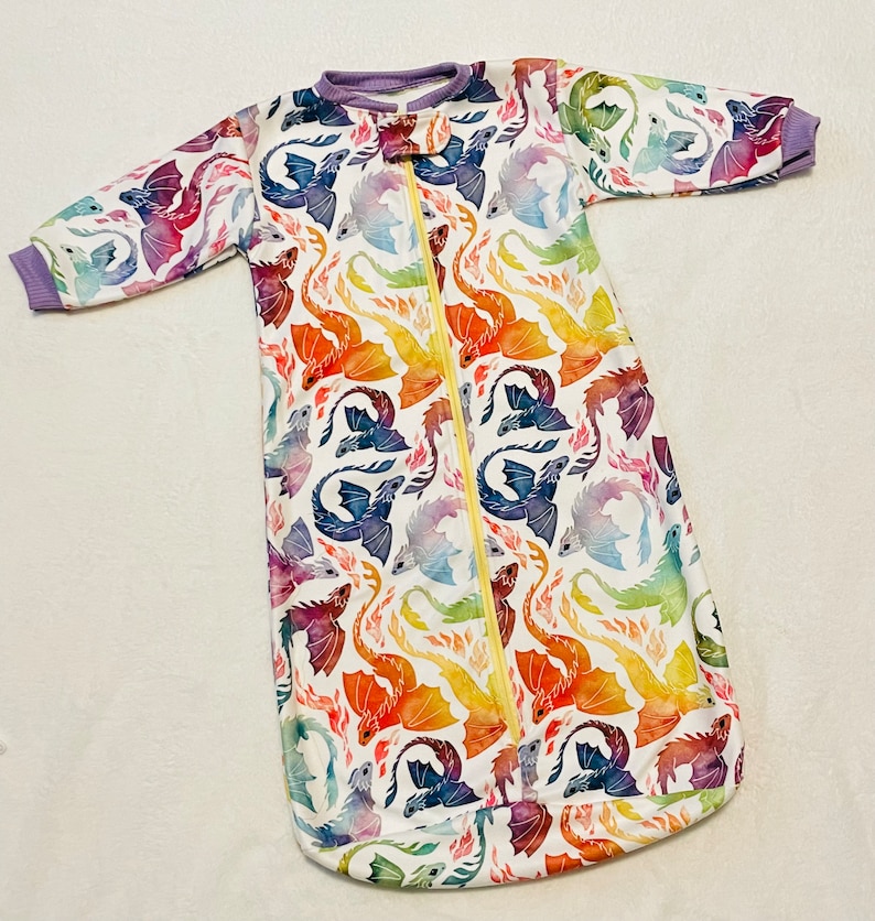 May include: A white baby sleeping bag with a zip fastening and a colourful watercolour dragon print. The dragons are in shades of blue, purple, orange, red and yellow.