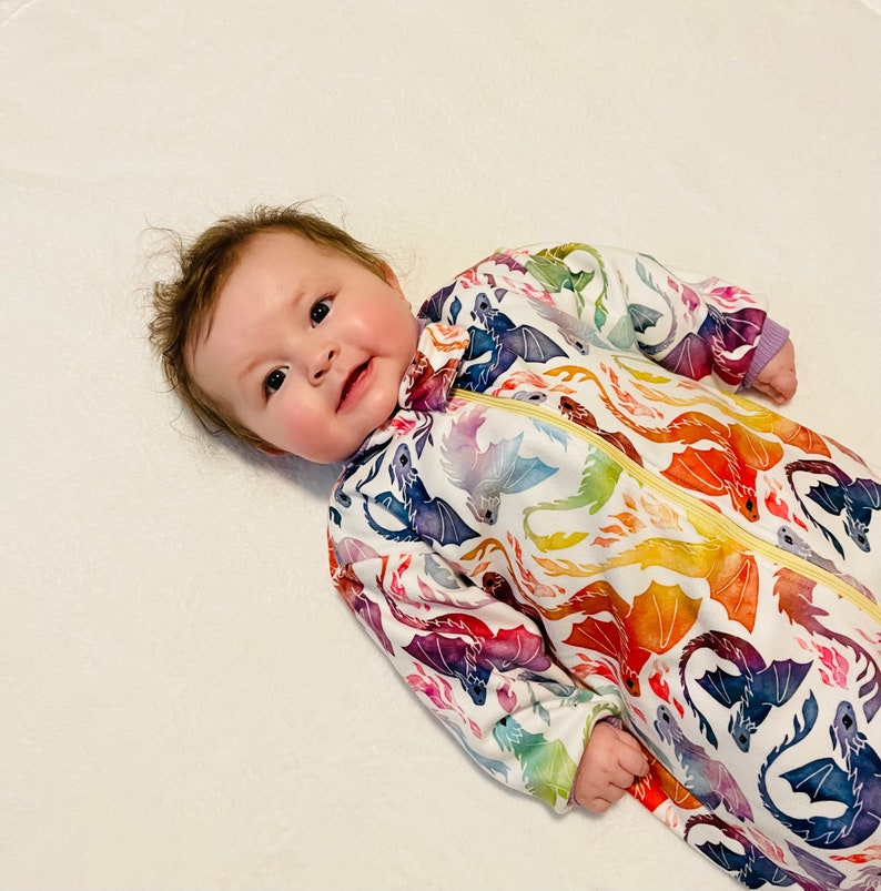May include: A baby wearing a white onesie with a colourful dragon print. The onesie has a zip down the front and is made of a soft, comfortable fabric.