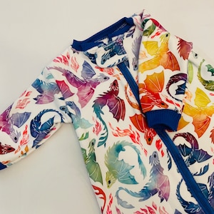 May include: A white baby onesie with a colourful watercolour dragon print. The onesie has a blue zipper and blue trim on the sleeves and neckline.