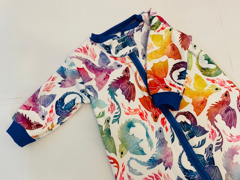 May include: A white baby onesie with a colourful watercolour dragon print. The onesie has a blue zipper and blue trim on the sleeves and neckline.