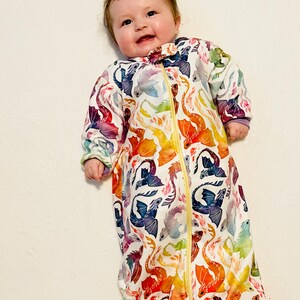 May include: A white baby sleeping bag with a colourful dragon print. The sleeping bag has a zipper fastening and is made from soft, breathable fabric.