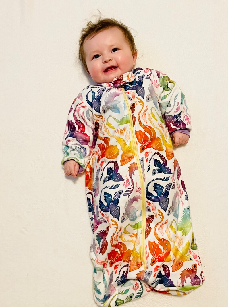 May include: A white baby sleeping bag with a colourful dragon print. The sleeping bag has a zipper fastening and is made from soft, breathable fabric.