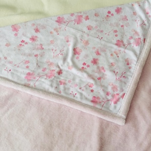 Puede incluir: Una manta rosa suave con un estampado floral blanco con flores rosas. La manta tiene una base de felpa rosa claro y un borde blanco. Una manta amarilla pálida es visible en el fondo.