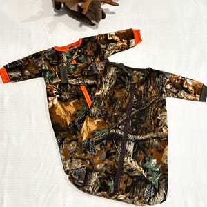 May include: Two baby sleep sacks with a camouflage pattern in shades of brown, green, and orange. One sleep sack has an orange trim and the other has a brown zipper. The sleep sacks are made of a soft material.