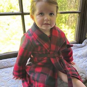 May include: A child wearing a red and black plaid robe with a tie belt. The robe is made of soft fleece and has a relaxed fit.