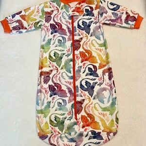 May include: A white sleep sack with a colourful dragon print. The sleep sack has a zipper down the front and long sleeves.