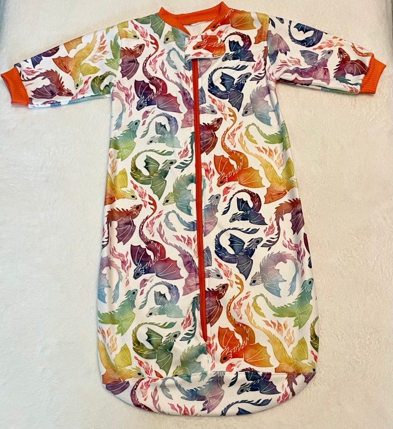 May include: A white sleep sack with a colourful dragon print. The sleep sack has a zipper down the front and long sleeves.