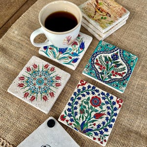 May include: A white ceramic mug filled with dark liquid sits on a floral coaster. Several square stone coasters with colorful floral designs are arranged around the mug. The coasters feature red, blue, and green patterns.