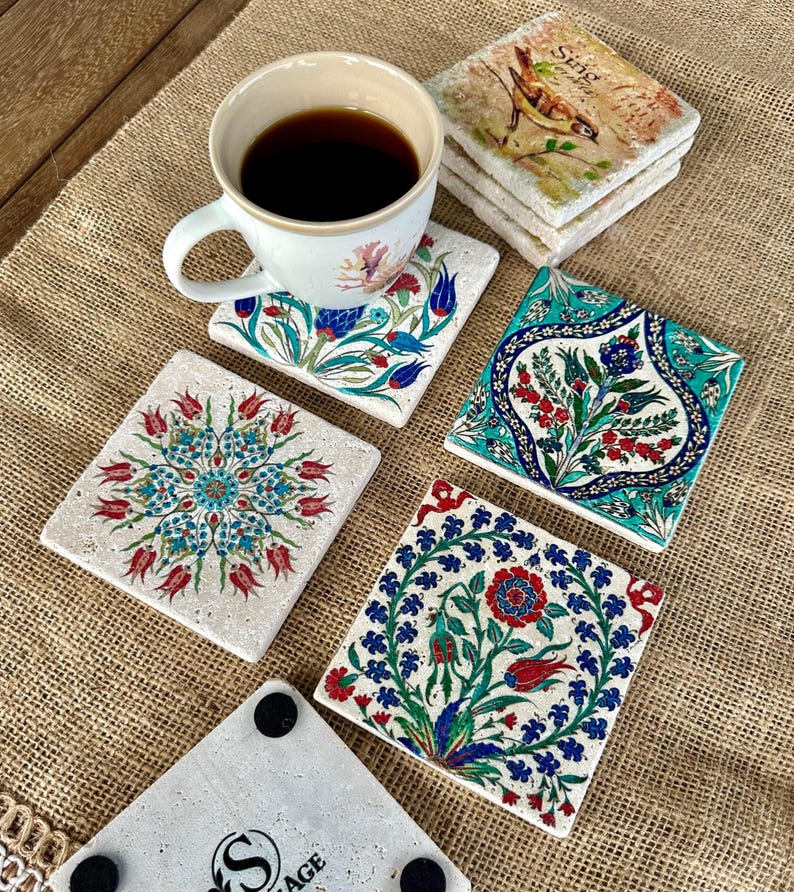 May include: A white ceramic mug filled with dark liquid sits on a floral coaster. Several square stone coasters with colorful floral designs are arranged around the mug. The coasters feature red, blue, and green patterns.