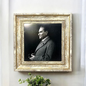 May include: A framed black and white portrait of a man in a tweed coat, set against a dark background. The portrait is enclosed in a distressed, ornate gold and cream-colored frame. The image is hanging on a light gray wall.