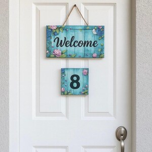 以下が含まれることがあります： ターコイズブルーの木製サインで、筆記体で「Welcome」と書かれ、ピンクのバラと青い花で飾られています。その下に、数字の「8」が表示された小さな正方形のサインがあります。どちらのサインも白いドアに掛けられています。