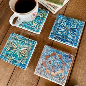 May include: A selection of square ceramic coasters featuring detailed floral and geometric patterns in blue, turquoise, and brown hues. A white coffee cup with a floral design rests on one coaster. The coasters are displayed on a wooden surface.