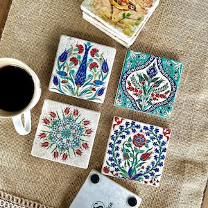 Puede incluir: Un conjunto de posavasos cuadrados de cerámica con intrincados diseños florales en rojo, azul y turquesa. Un posavasos presenta una ilustración de un pájaro. Una taza de café está cerca.