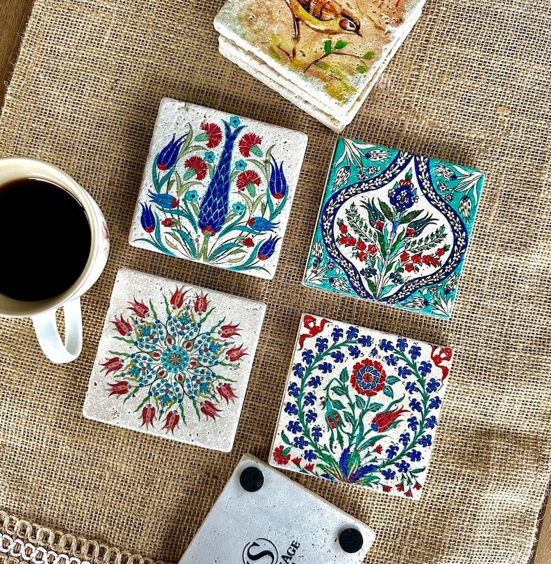 May include: A collection of square ceramic coasters with intricate floral designs in red, blue, and turquoise. One coaster features a bird illustration. A cup of coffee sits nearby on a burlap surface.